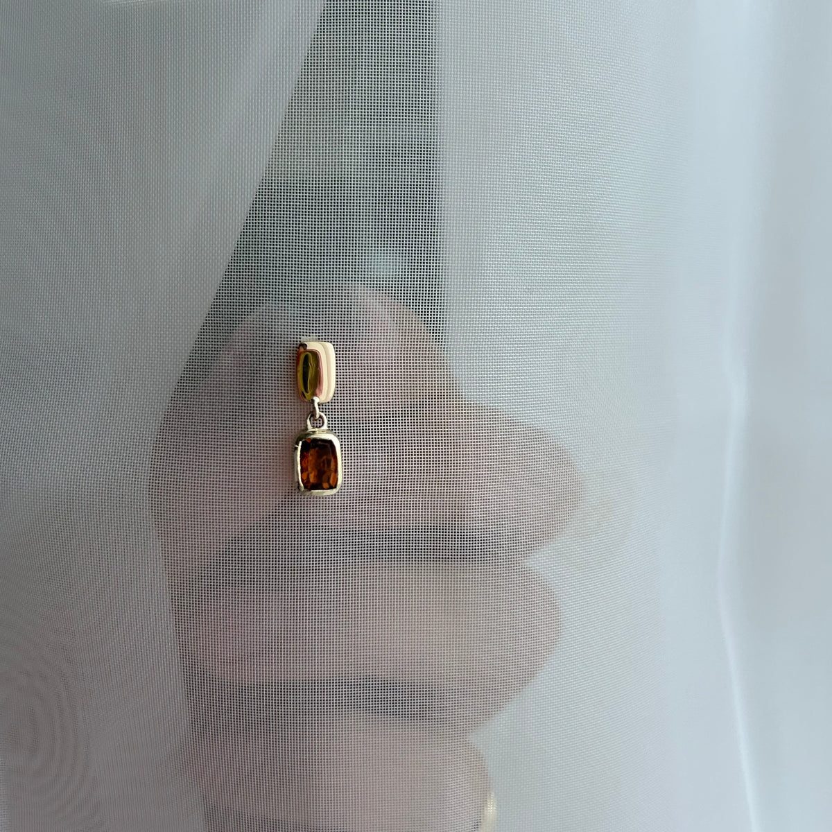 A close-up of a hand holding a pair of gold drop earrings with rectangular amber-brown gemstones behind a sheer white curtain.
