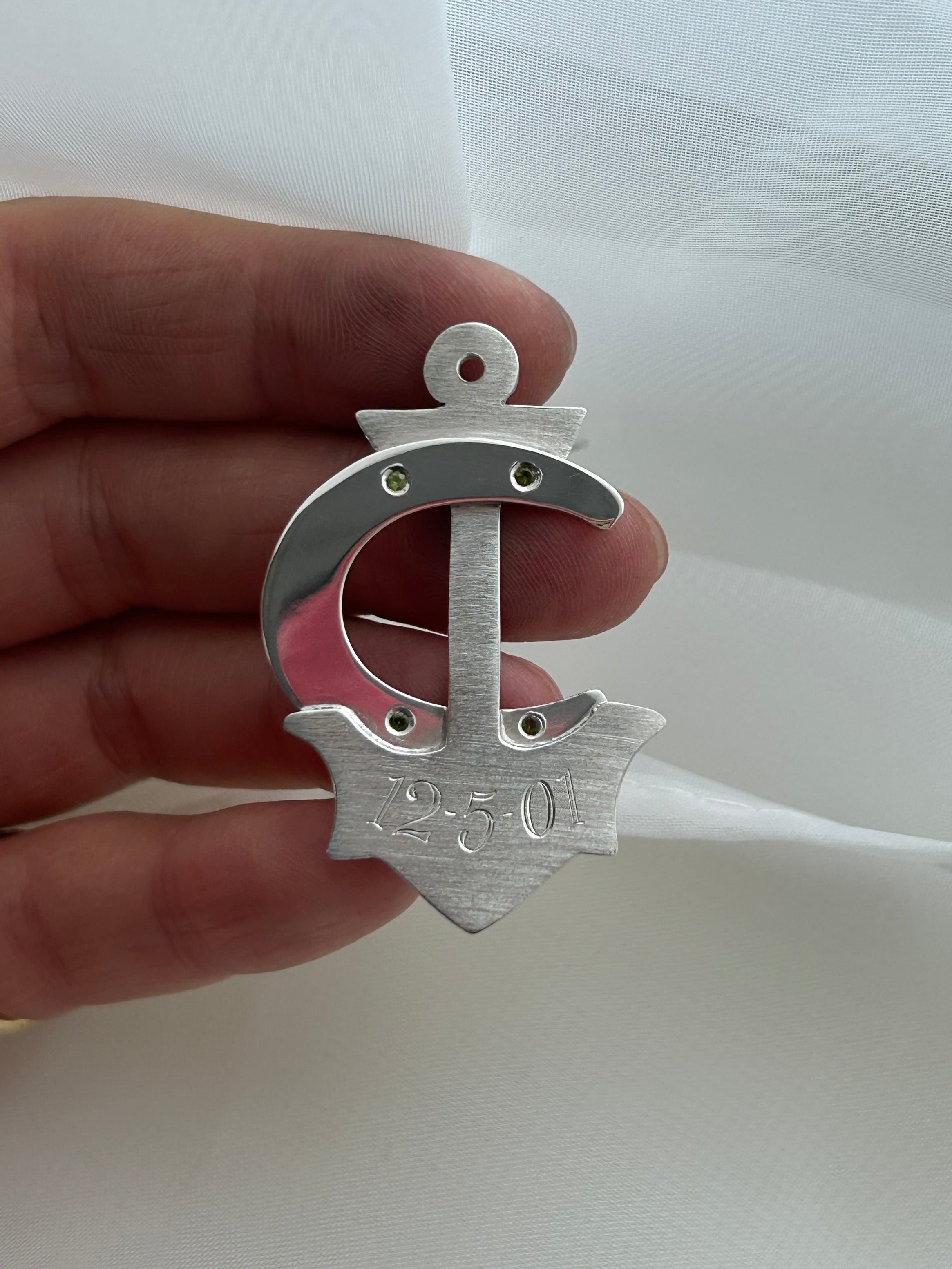 A hand holding a silver anchor-shaped pendant or charm against a white fabric background.