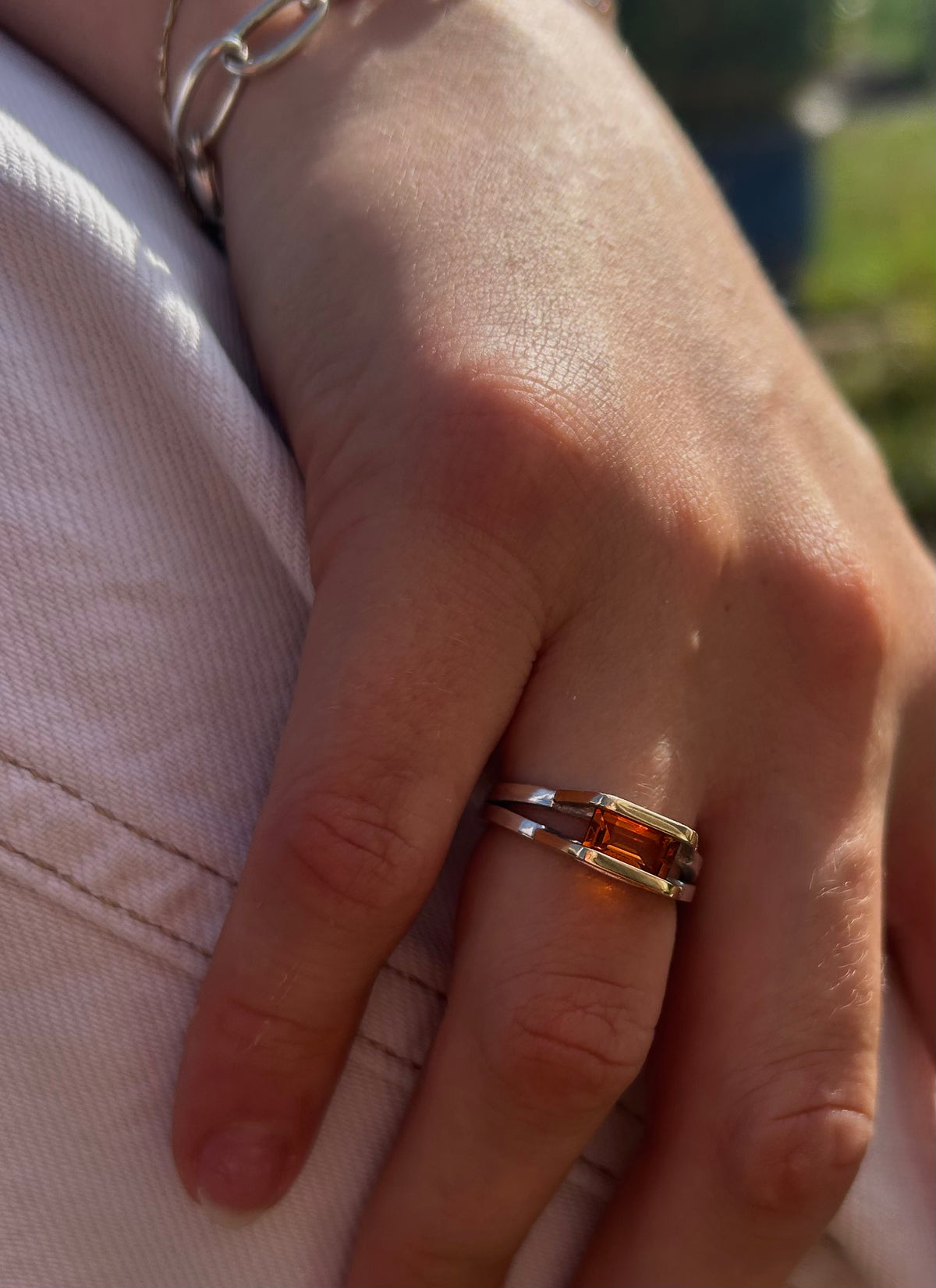 A close-up of a person's hand wearing a silver ring with a rectangular amber-orange stone, resting on light-colored denim fabric.