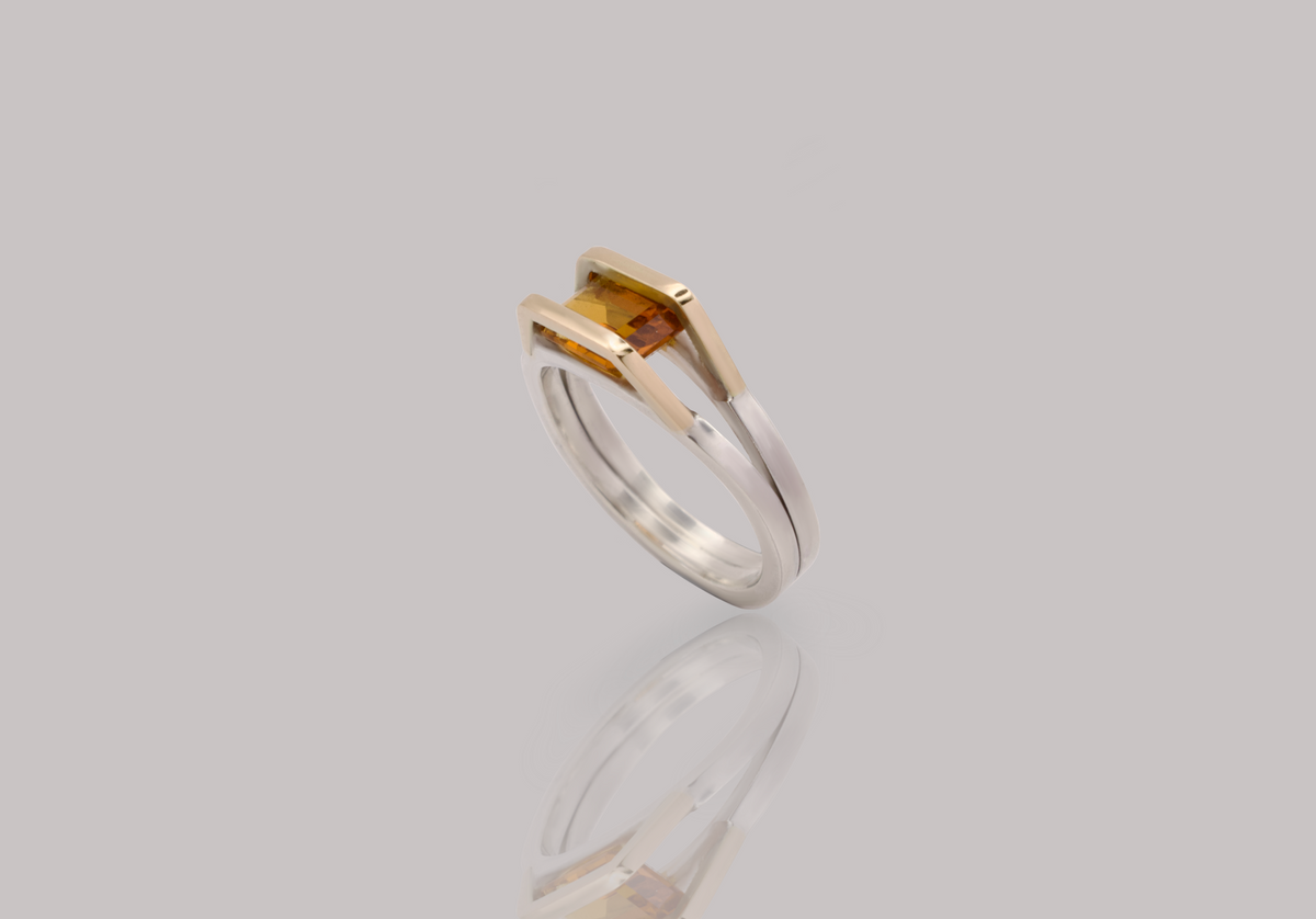 A two-tone ring featuring a rectangular yellow gemstone in a prong setting with one gold-toned half and one silver-toned half, shown on a light gray reflective background.