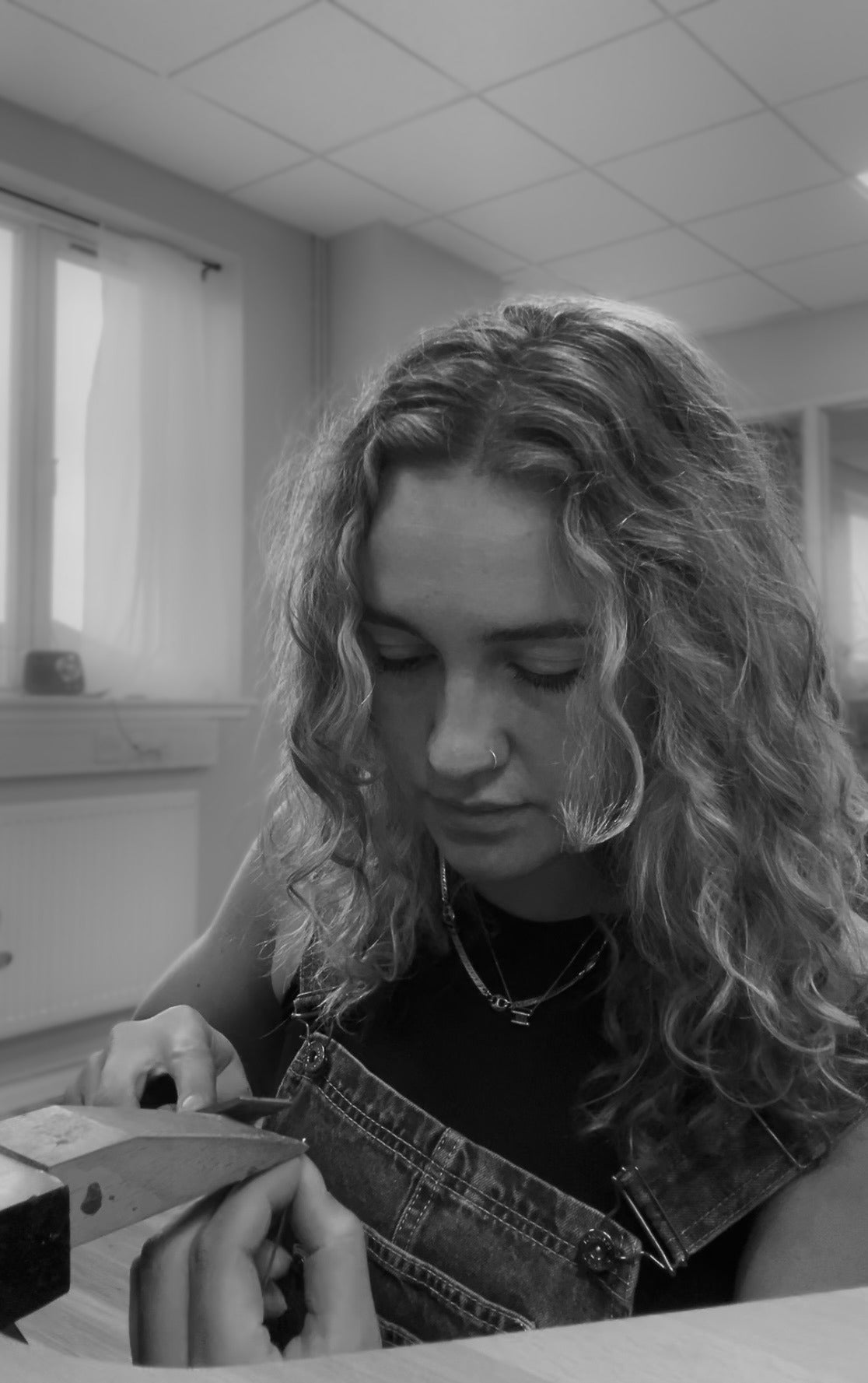 A black and white lifestyle photograph of a woman with long curly hair looking down while wearing denim overalls over a black top in an indoor setting.