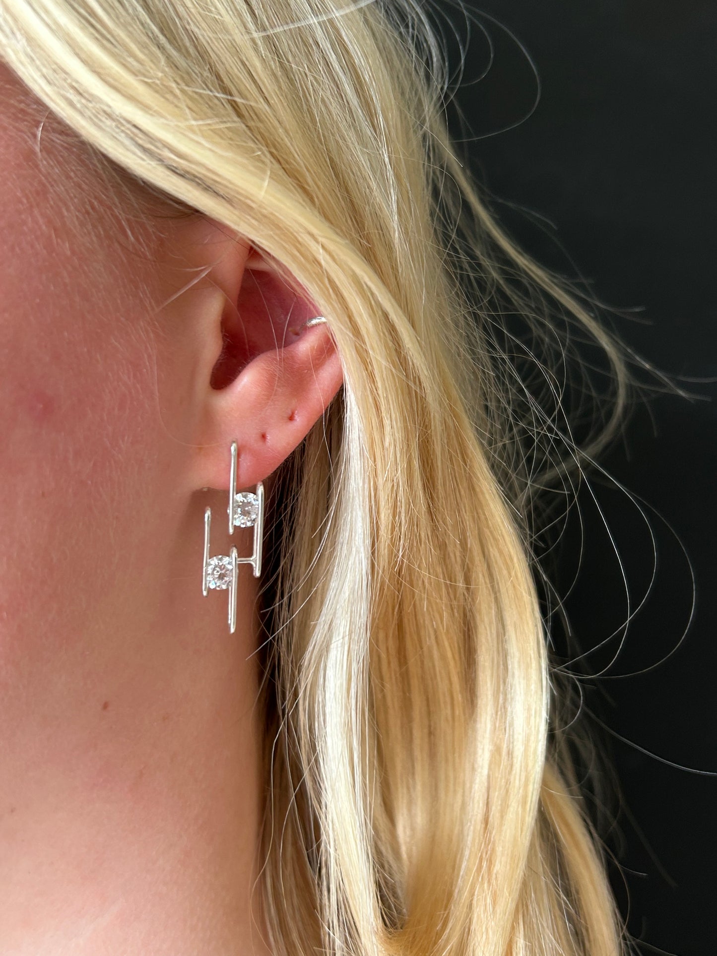 Close-up of an ear wearing a silver earring with two small stones, with blonde hair in the foreground.