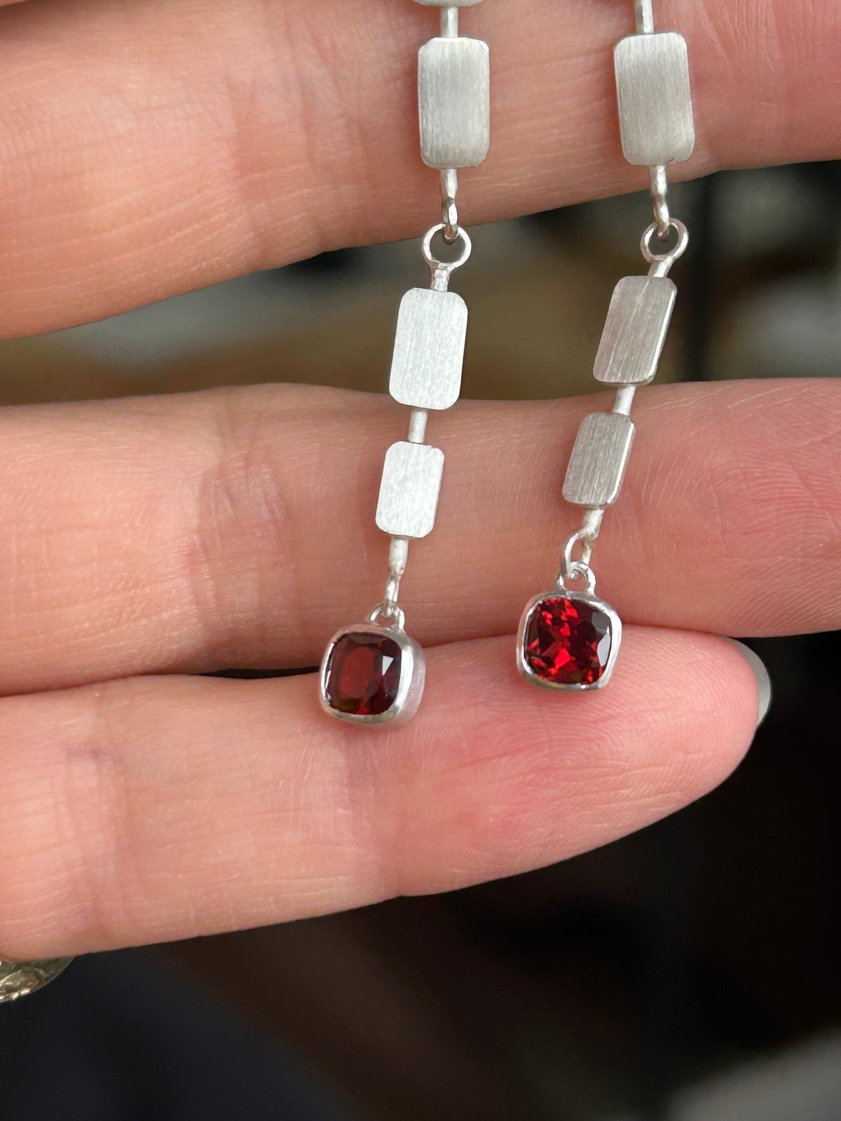 A close-up of a hand holding a pair of silver drop earrings with rectangular brushed metal beads and square-cut red gemstones.