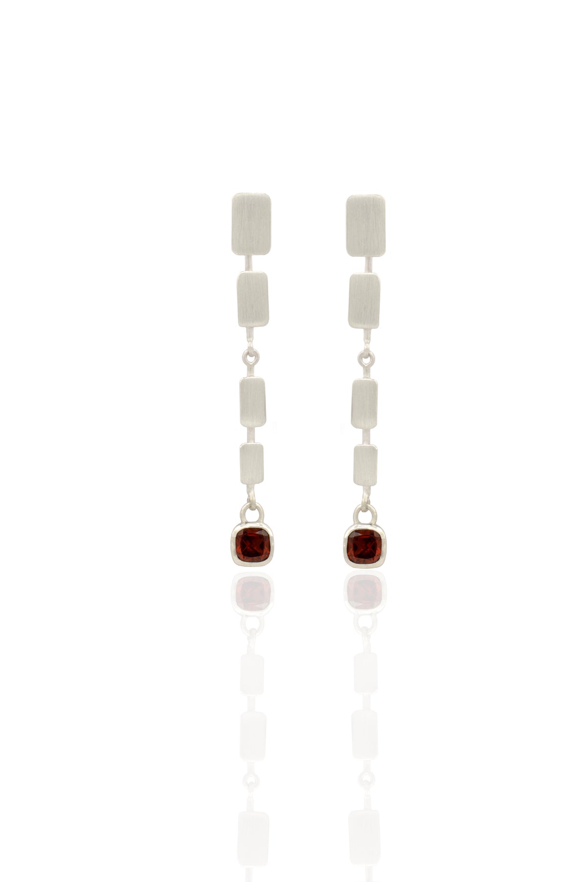 A pair of silver drop earrings featuring a vertical stack of textured rectangular silver beads, connected by small silver links, with each earring ending in a bezel-set round dark red gemstone, displayed on a clean white reflective surface.