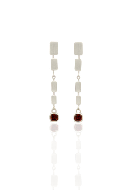 A pair of silver drop earrings featuring a vertical stack of textured rectangular silver beads, connected by small silver links, with each earring ending in a bezel-set round dark red gemstone, displayed on a clean white reflective surface.
