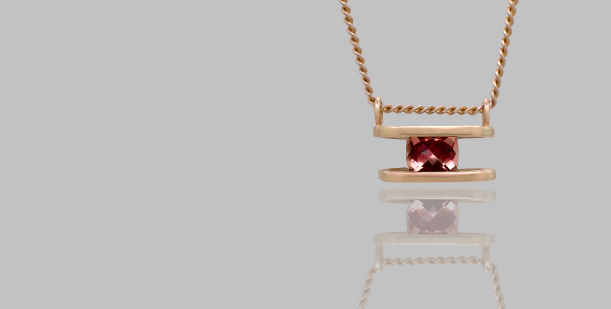 A rose gold necklace with a minimalist pendant featuring a central faceted burgundy gemstone, displayed on a solid gray background with a soft reflection below.