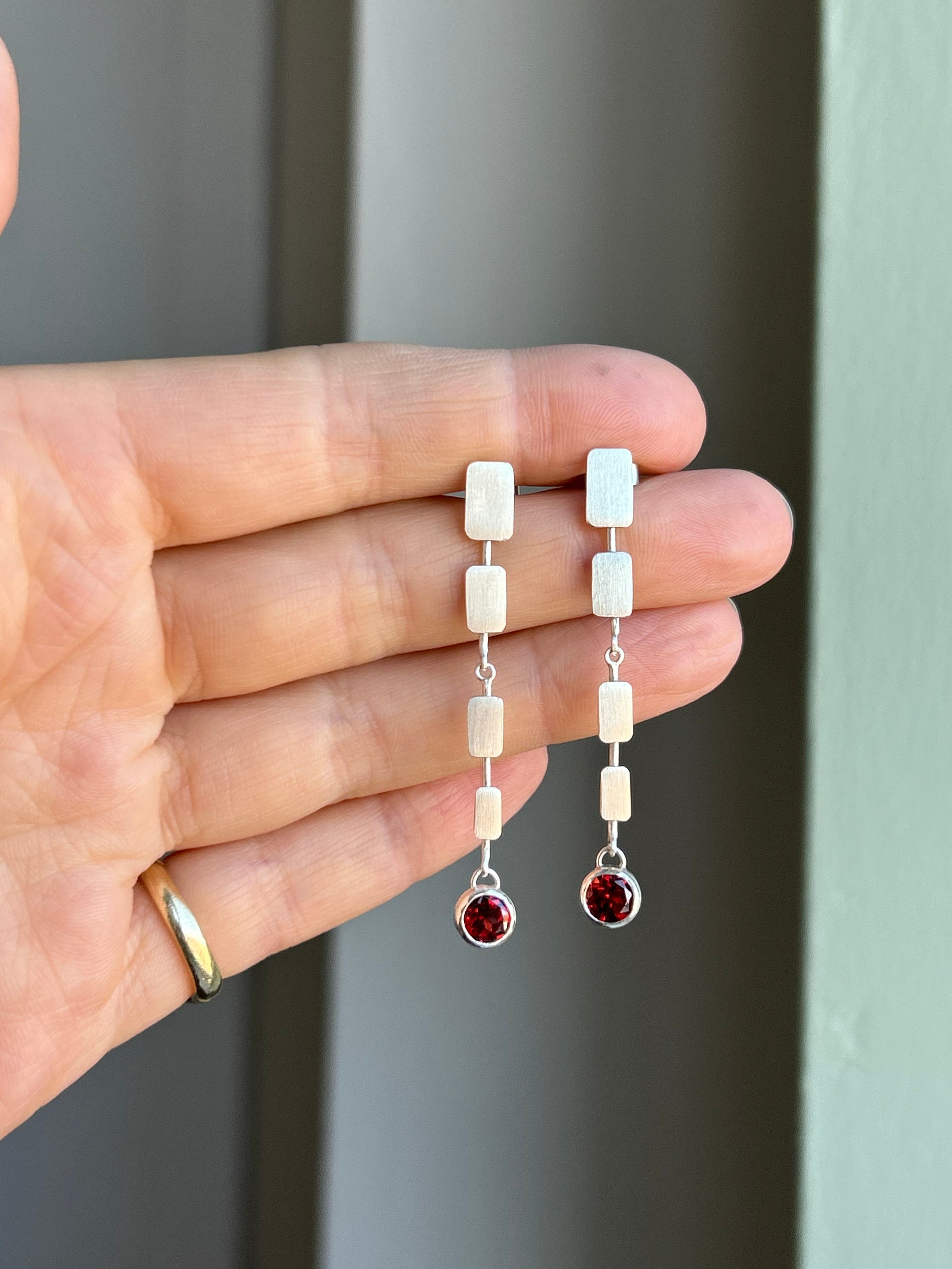 A person holding a pair of silver drop earrings with garnet stones.