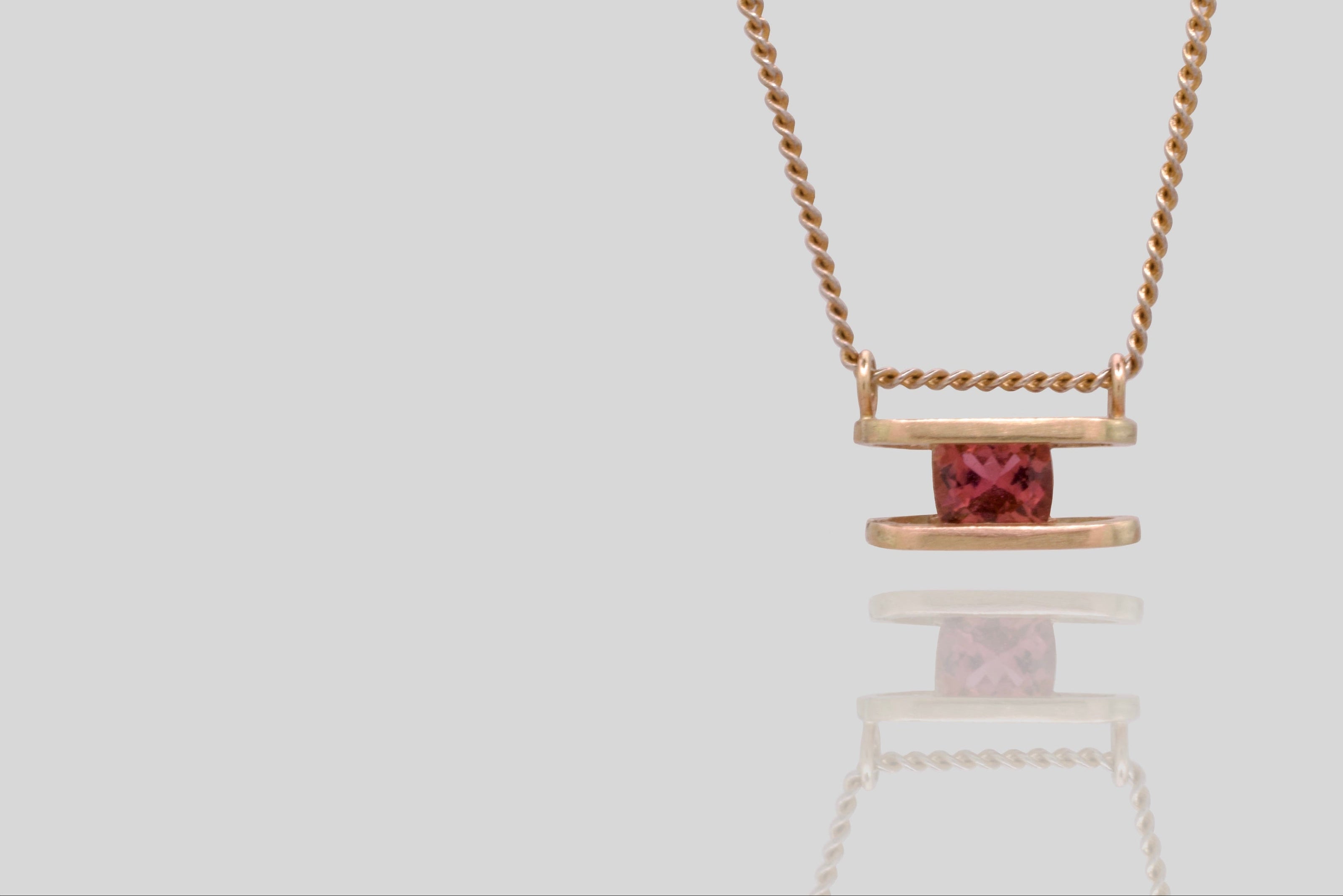 Gold necklace with a dark pink gemstone pendant on a light grey background