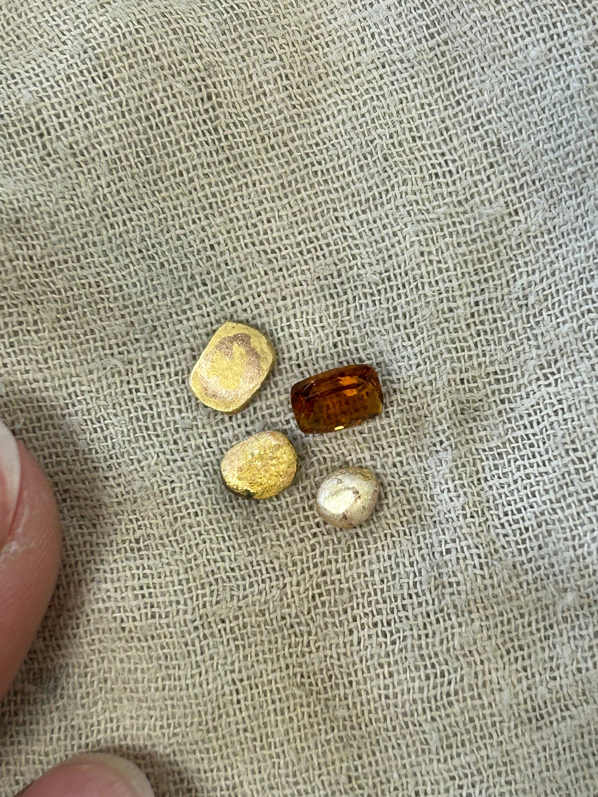 Four small polished gemstones or beads in yellow, cream, and amber-brown tones arranged on a beige woven fabric, with a person's fingers visible at the bottom left for scale.