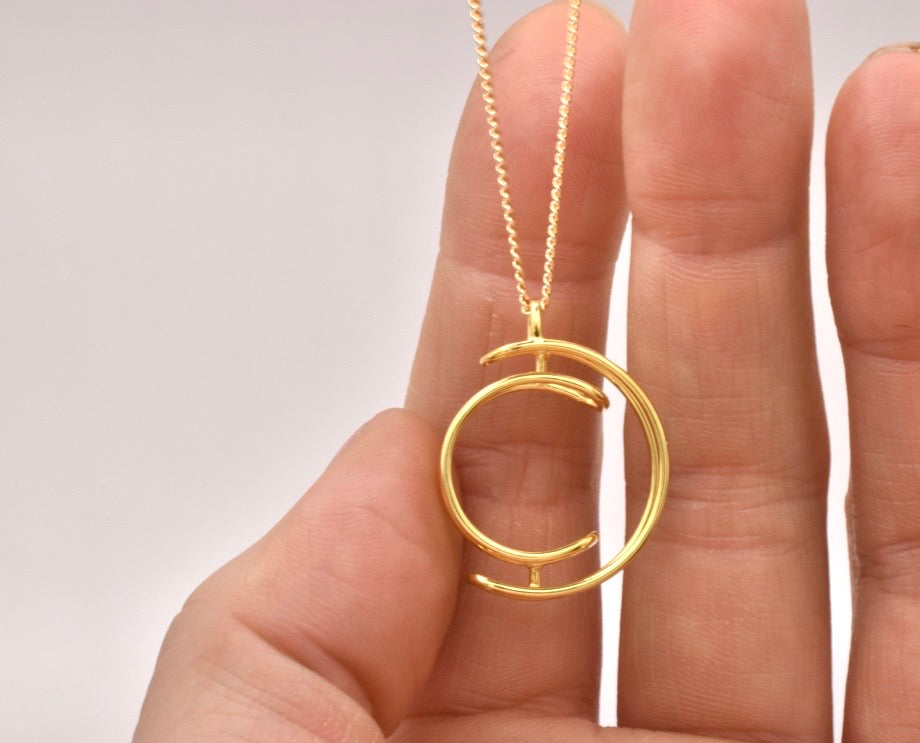 Gold necklace with a circular pendant held between fingers against a neutral background