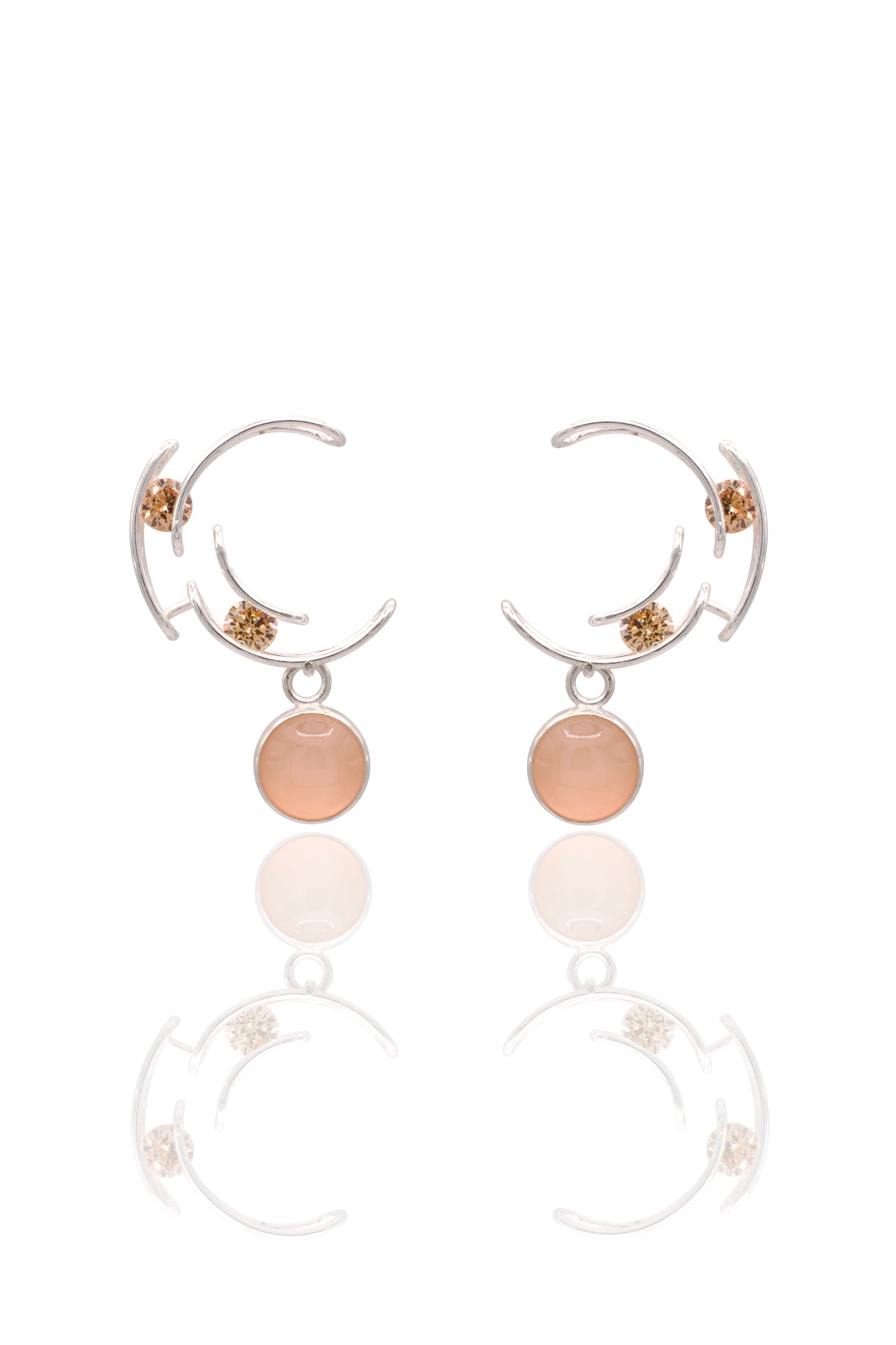 A pair of silver crescent-shaped hoop earrings with small gold floral accents and a round peach-pink stone dangle, shown on a white background with a soft reflection below.