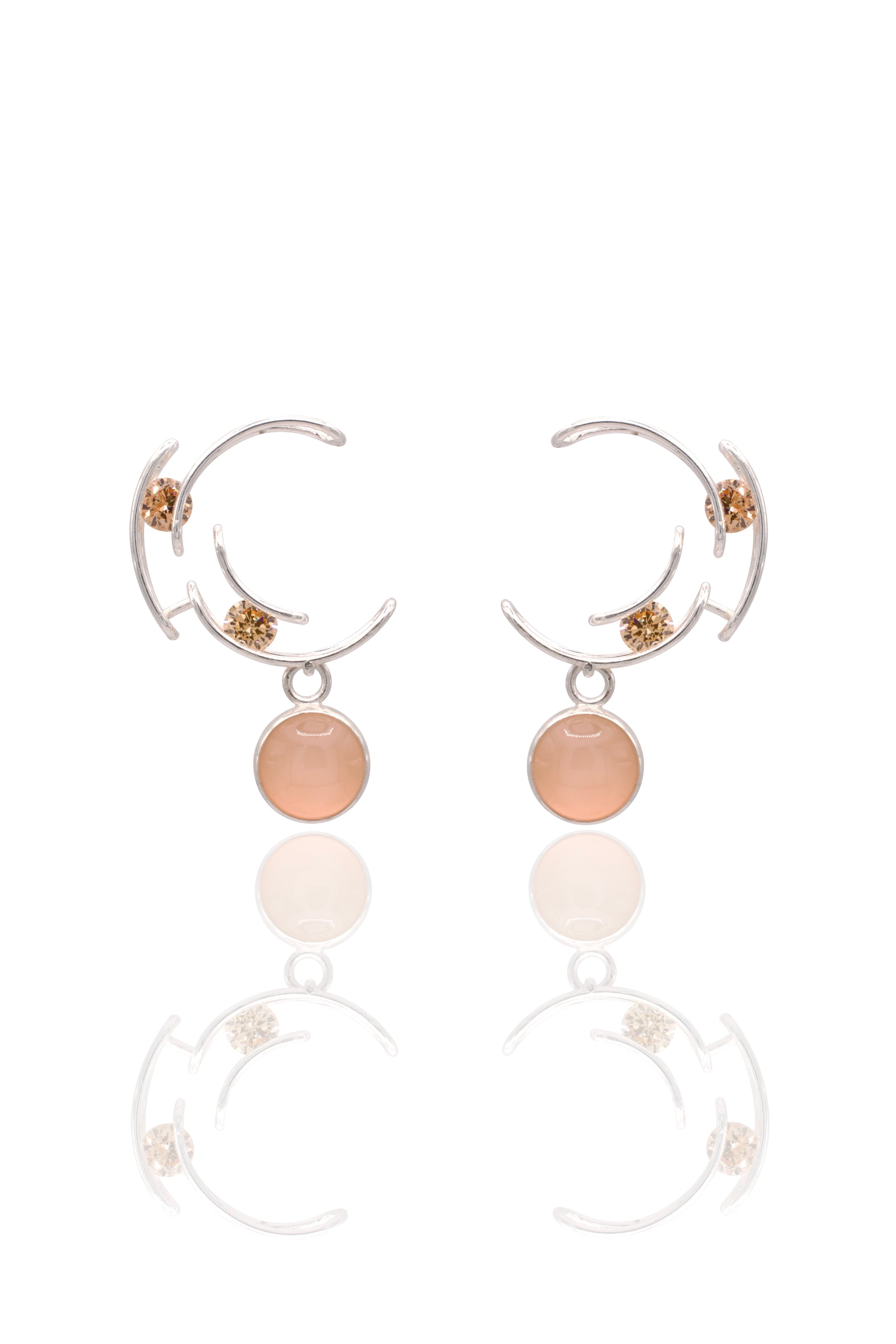 A pair of silver crescent-shaped hoop earrings with small gold floral accents and a round peach-pink stone dangle, shown on a white background with a soft reflection below.