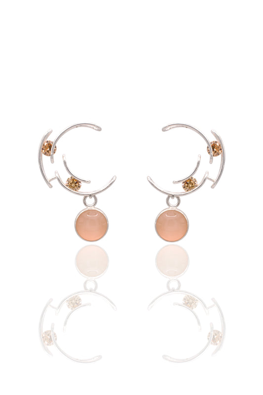 A pair of silver crescent-shaped hoop earrings with small gold floral accents and a round peach-pink stone dangle, shown on a white background with a soft reflection below.