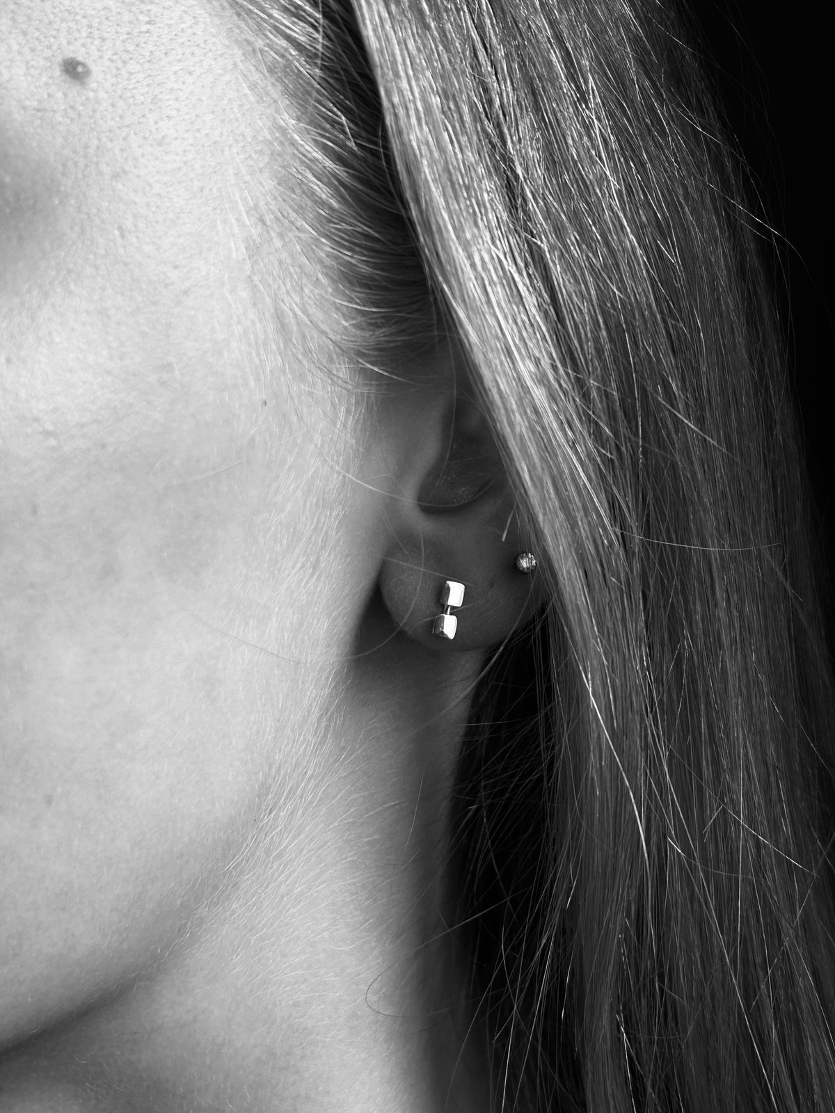 Close-up of a person's ear wearing a small silver geometric stud earring and a tiny round stud, shown in black and white with long hair visible.