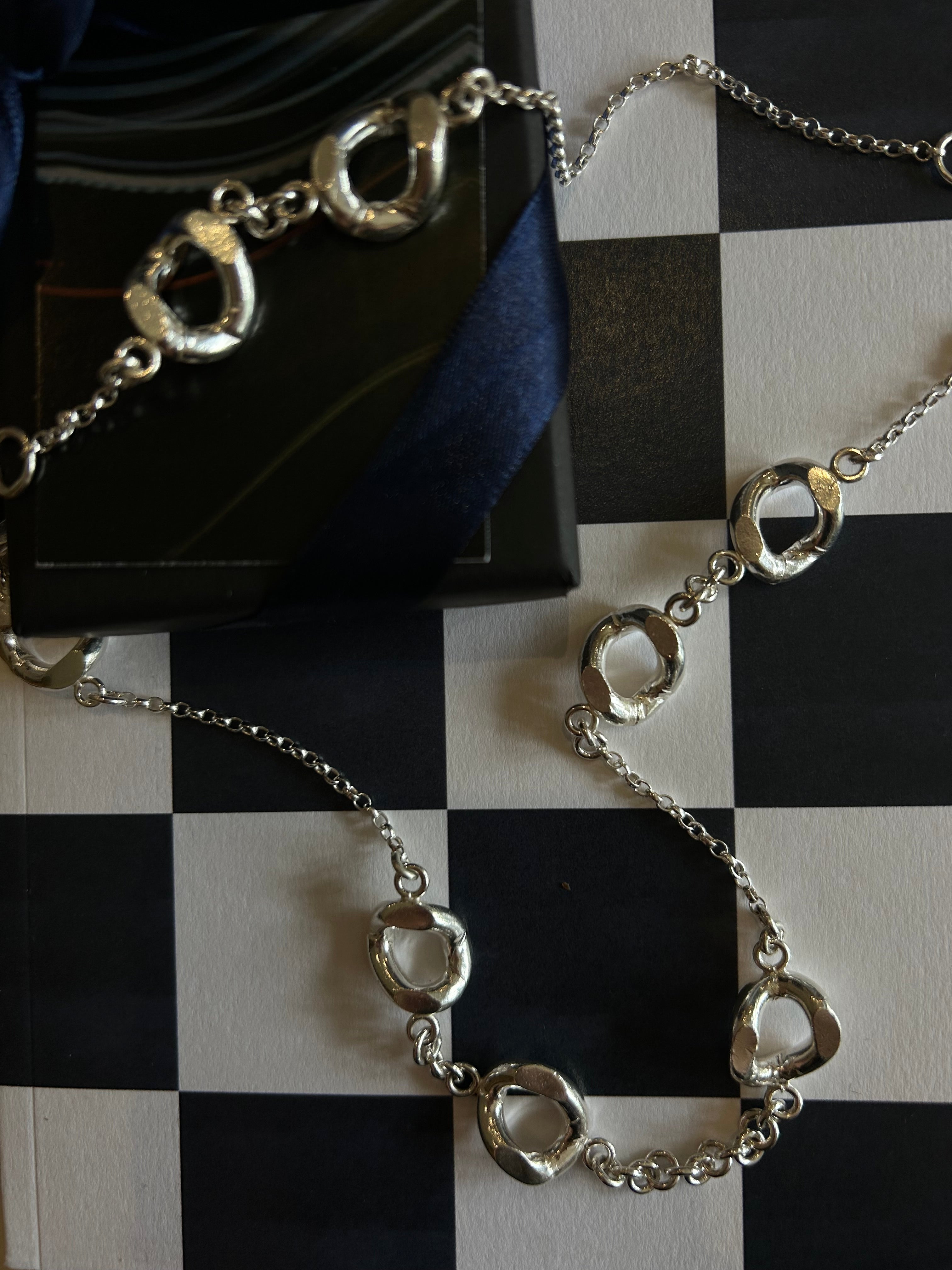 A silver-toned chain necklace with irregular, textured circular links, displayed on a black-and-white checkered surface next to a dark box and navy ribbon.