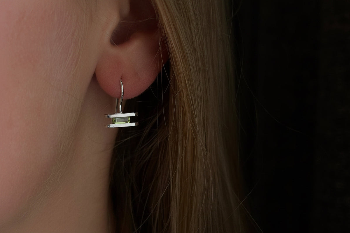 A close-up of a person's ear wearing a small silver geometric dangle earring against dark hair and a dark background.