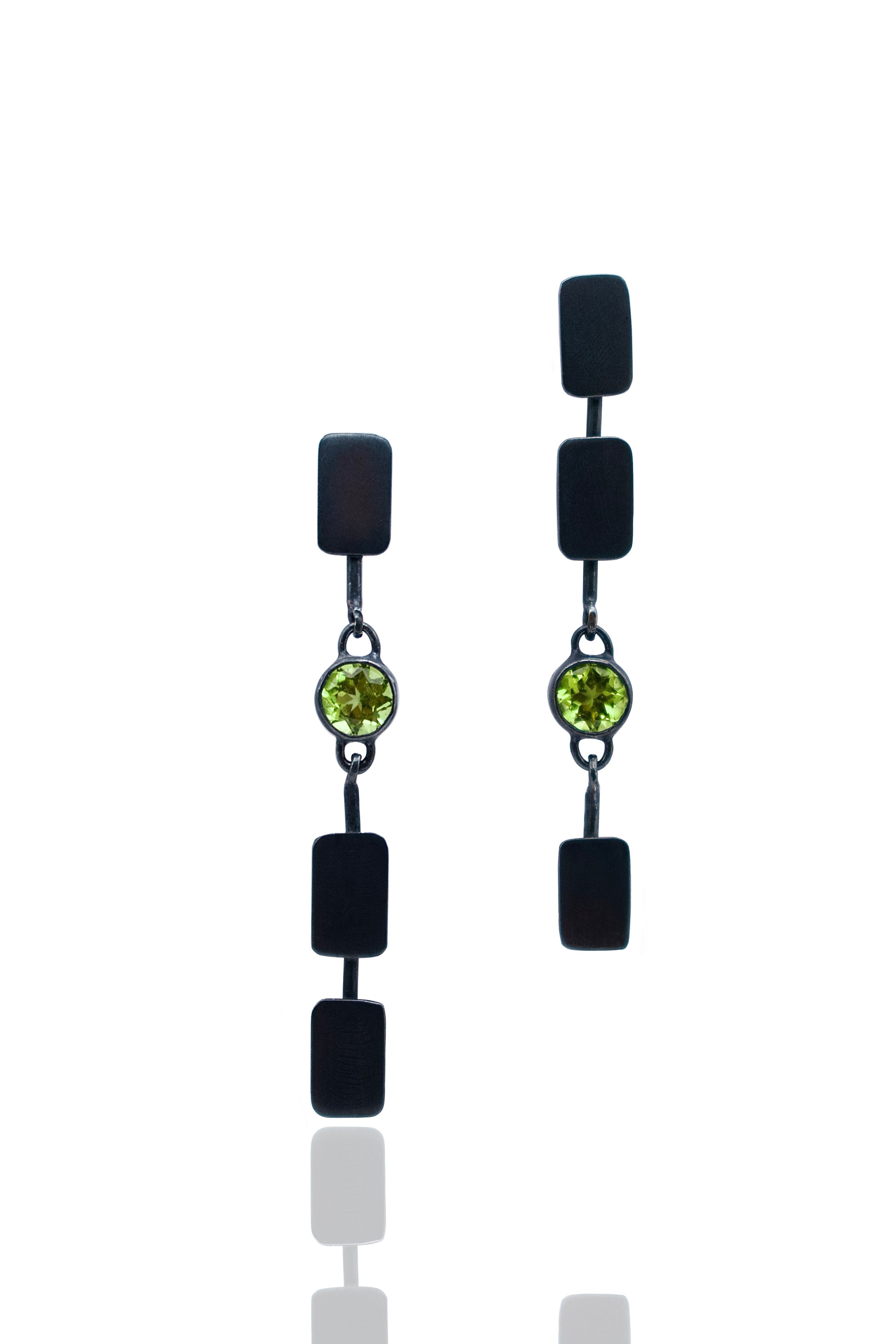 A pair of black drop earrings with rectangular black beads, a small round green gemstone in the center, and silver-toned chain links, shown on a white background with a faint reflection below.