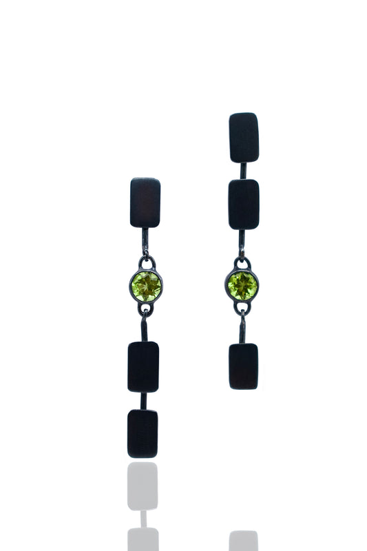 A pair of black drop earrings with rectangular black beads, a small round green gemstone in the center, and silver-toned chain links, shown on a white background with a faint reflection below.