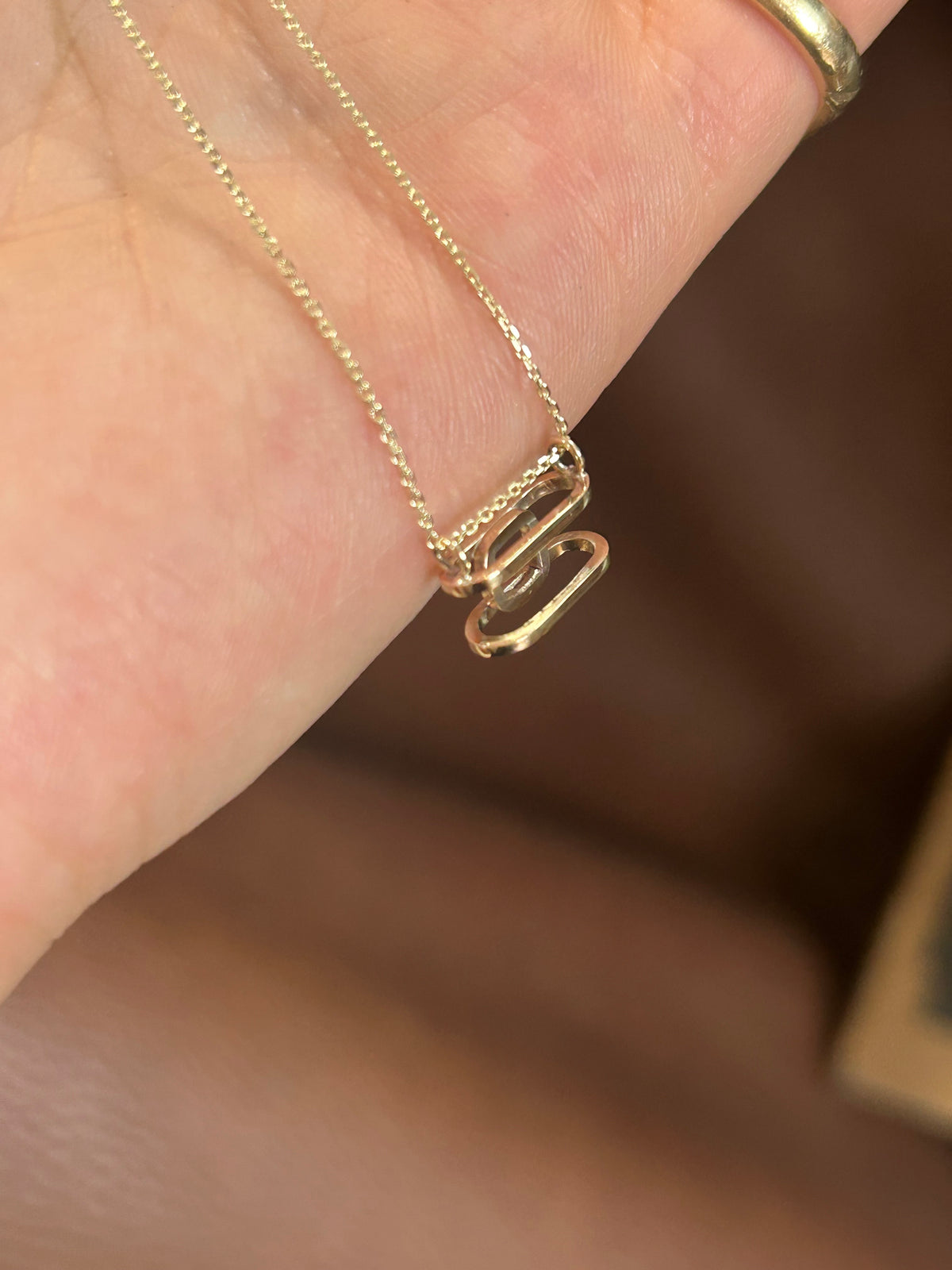 A close-up of a delicate gold chain necklace with a small interlocking double-loop pendant, worn on a person's finger.